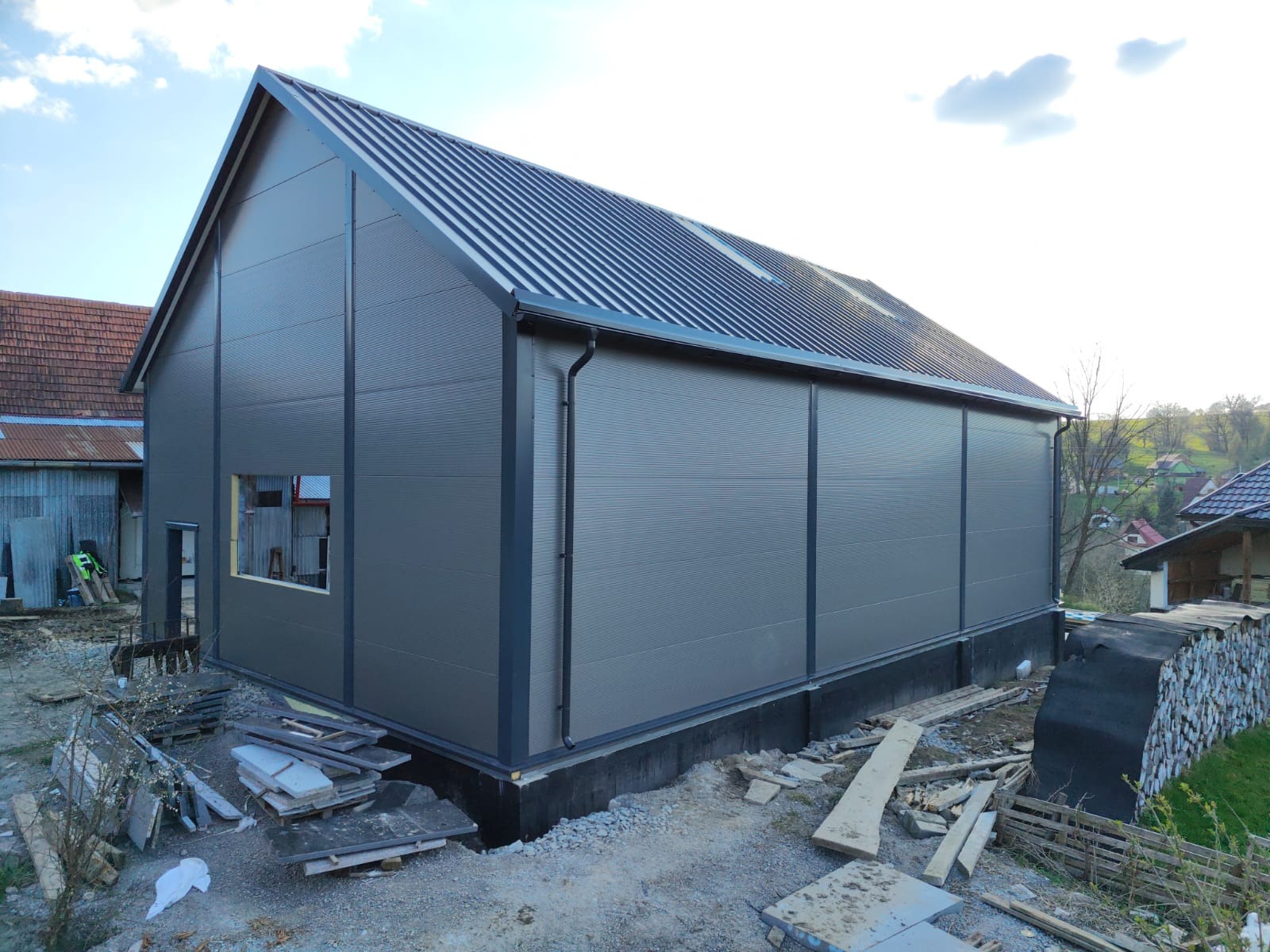 Hala z szarej płyty warstwowej z oknem i dachem z blachy trapezowej, w trakcie budowy. Widoczne materiały budowlane i elementy konstrukcyjne.