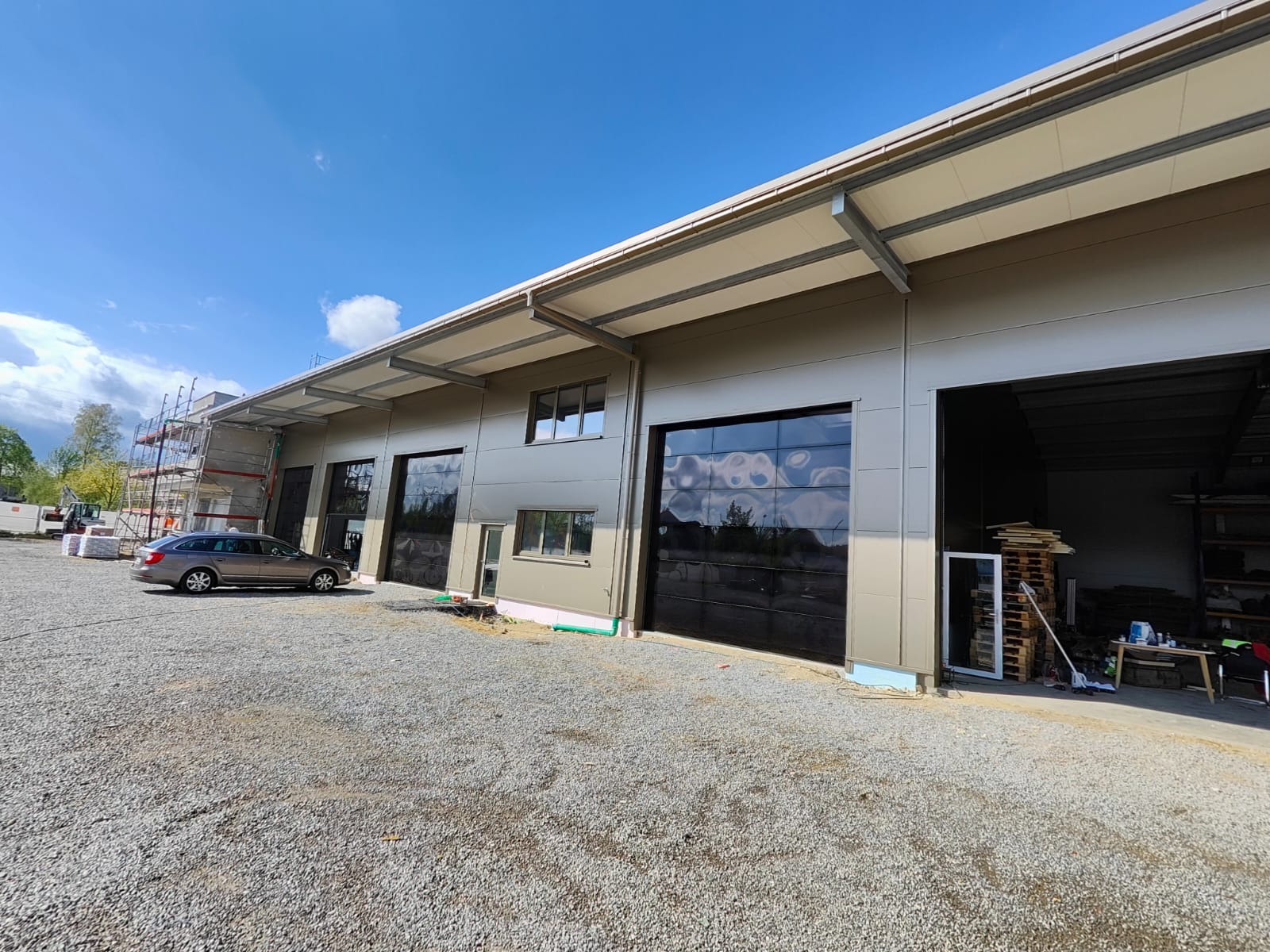 Nowoczesna hala z płyt warstwowych w trakcie budowy, widoczne szklane bramy segmentowe, szare elewacje i rusztowania. Na pierwszym planie szary samochód osobowy.