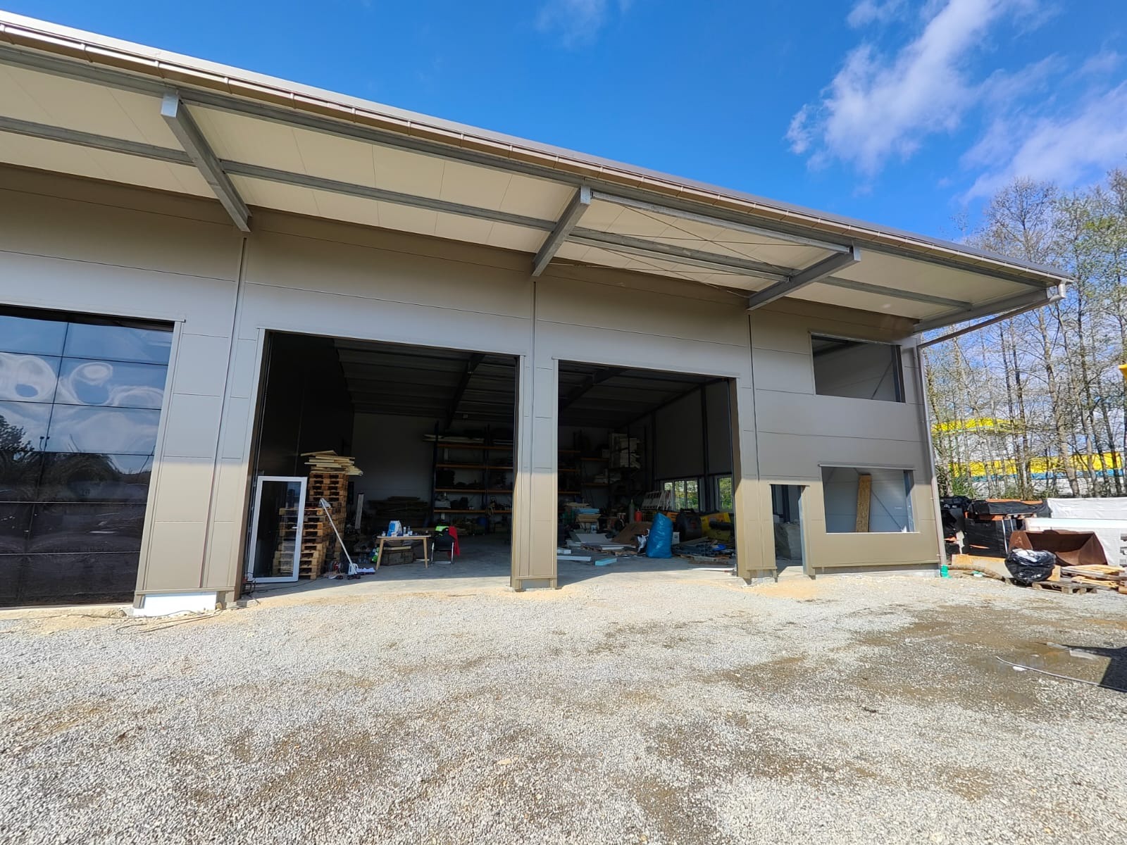 Nowoczesna hala z płyt warstwowych w trakcie budowy, widoczne otwarte wrota i elementy konstrukcyjne. Jasne niebo z chmurami w tle.