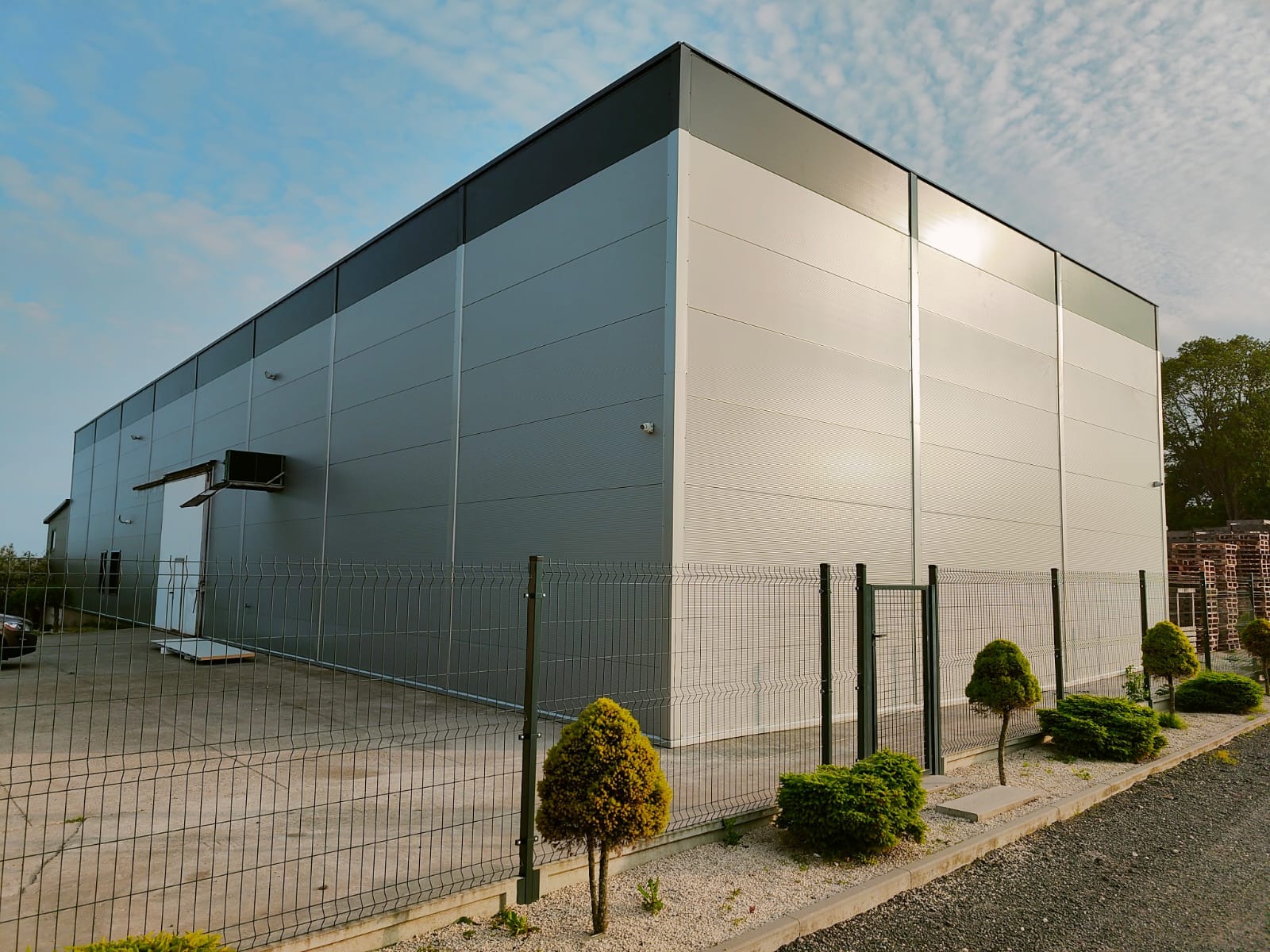 Nowoczesna hala magazynowa z płyt warstwowych, widoczna zza ogrodzenia panelowego. Jasna elewacja odbija światło słoneczne. Widoczne drzwi i fragment zieleni.