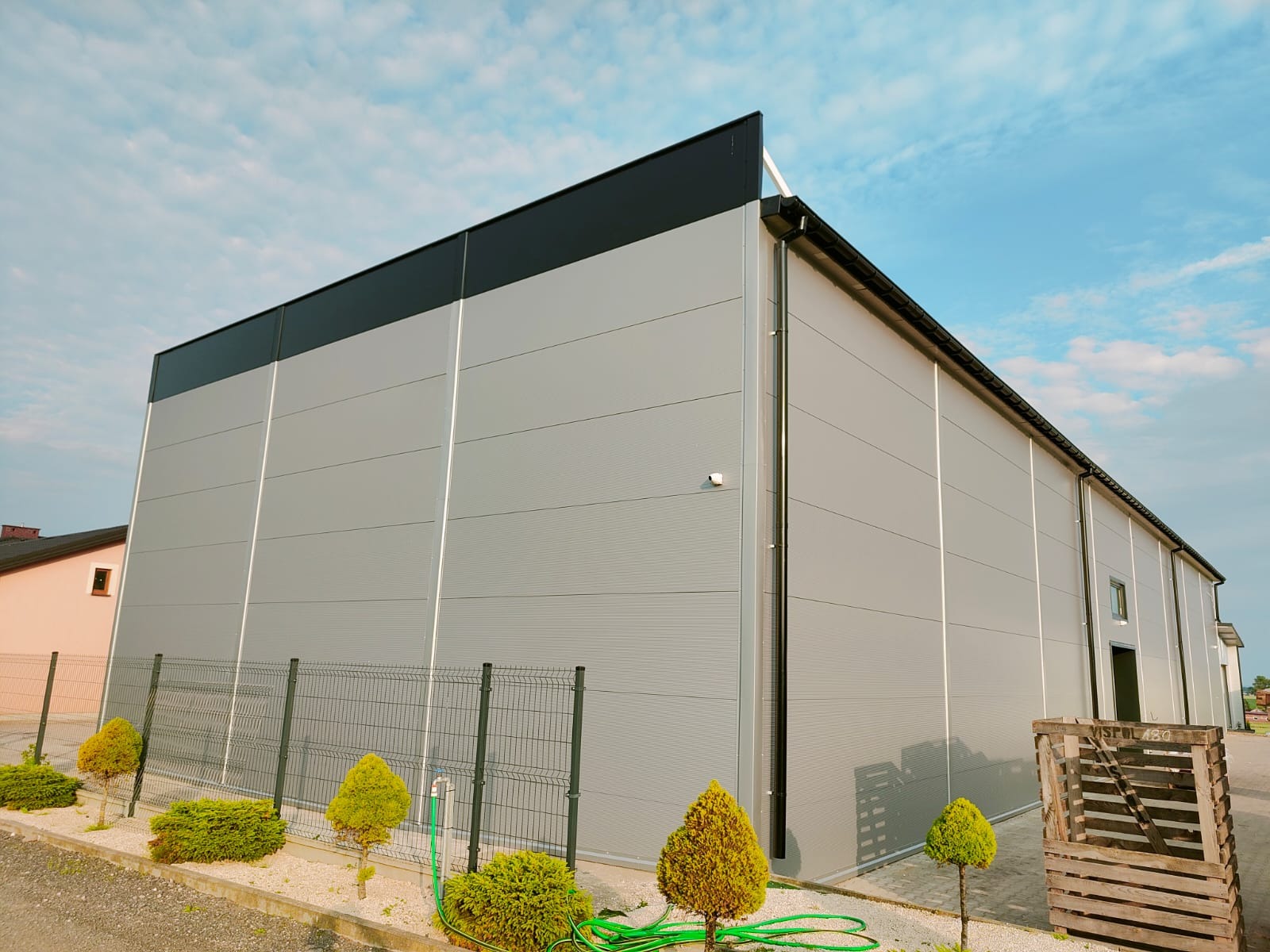 Nowoczesna hala z szarej płyty warstwowej, czarny dach, ogrodzenie z zielonymi krzewami. Widok z zewnątrz na tle błękitnego nieba.