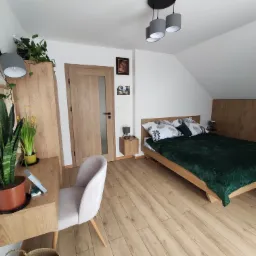 Pokój na poddaszu z drewnianą podłogą, łóżkiem z zieloną narzutą, biurkiem z szafką i roślinami doniczkowymi, oraz krzesłem obitym jasnym materiałem. Na ścianie wiszą obrazy i lampa.