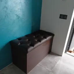 Tapicerowana pufa w ciemnobrązowej skórze, z pikowaniem w romby, stojąca na szarych płytkach pod ścianą w kolorze turkusowym i białą ścianą z włącznikami światła.