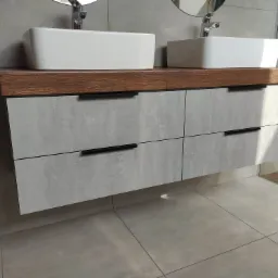 Łazienkowa szafka pod umywalki nablatowe z drewnianym blatem i szarymi frontami szuflad z czarnymi, podłużnymi uchwytami, umieszczona na tle szarej ściany i podłogi z płytek.