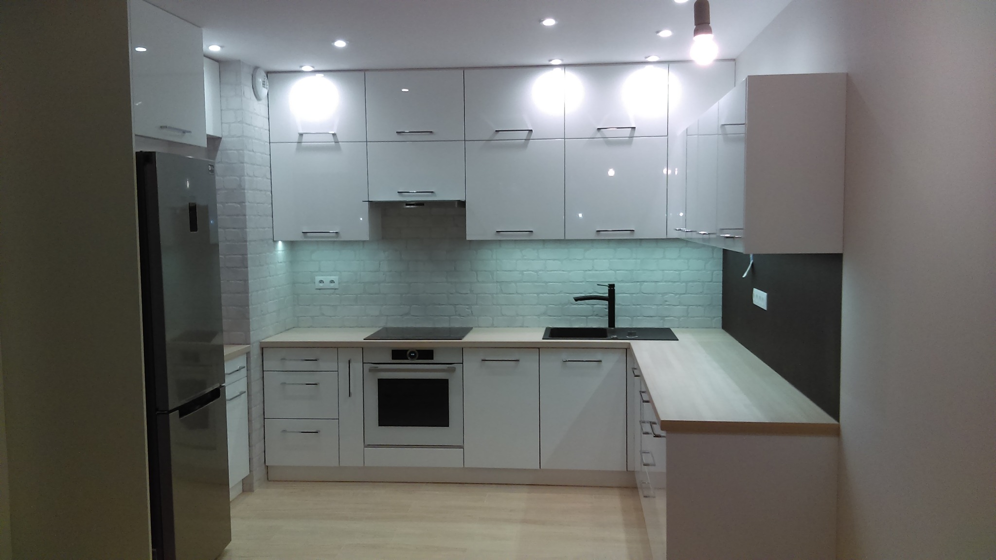 Nowoczesna, biała kuchnia z błyszczącymi frontami, oświetleniem punktowym i ceglaną ścianą w tle, uzupełniona o srebrną lodówkę i drewniany blat.