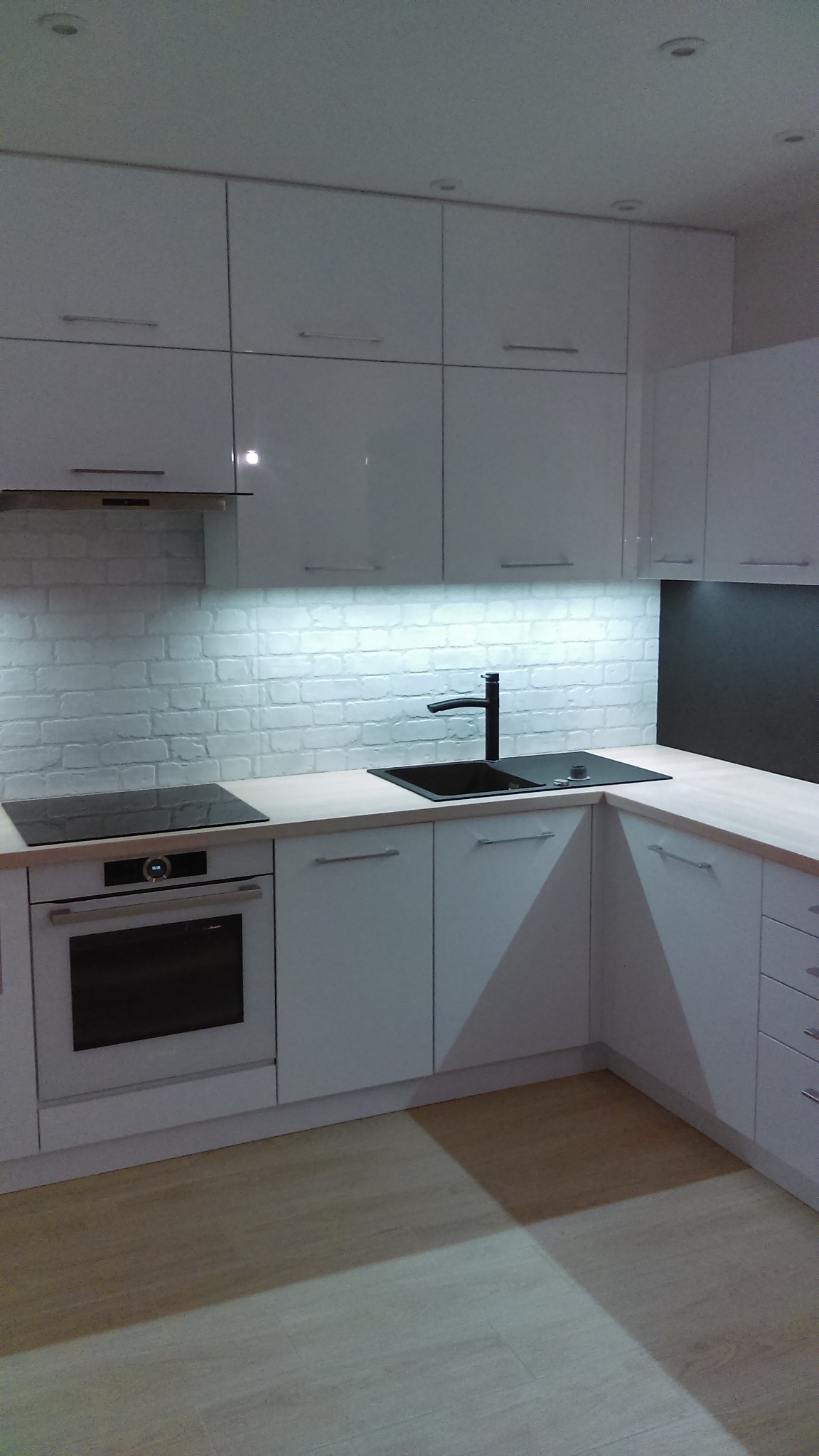 Nowoczesna, biała kuchnia z błyszczącymi frontami szafek, czarną armaturą i płytą indukcyjną, oświetlona listwą LED nad blatem wykończonym w jasnym drewnie.