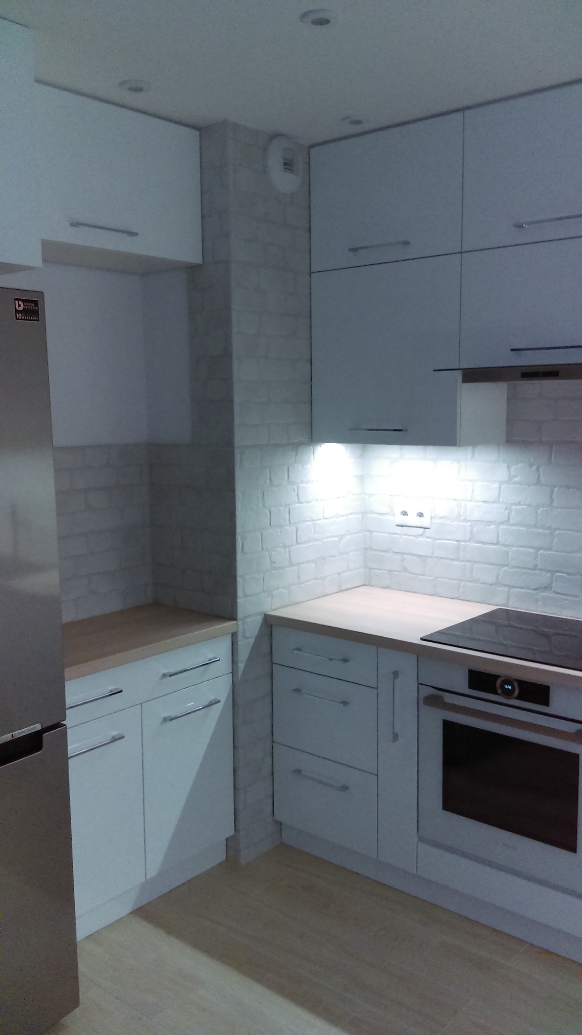 Nowoczesna, jasna kuchnia z białymi frontami szafek i blatem z jasnego drewna, oświetlona punktowo, z białymi cegiełkami na ścianie i wbudowanym piekarnikiem.