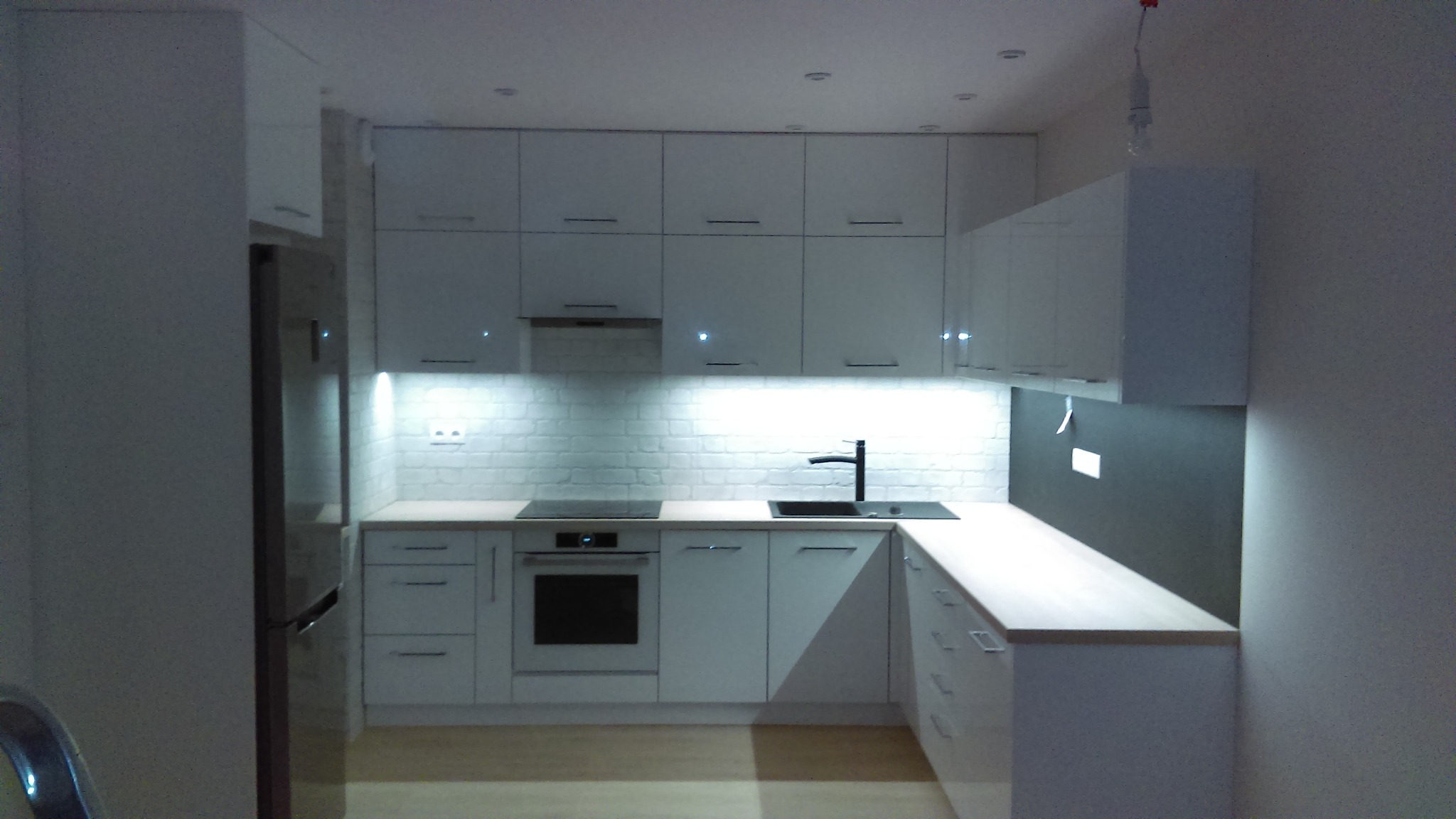 Nowoczesna biała kuchnia z białymi, lakierowanymi frontami, podświetleniem LED pod szafkami wiszącymi i blatem w kolorze jasnego drewna, czarny zlew i bateria.