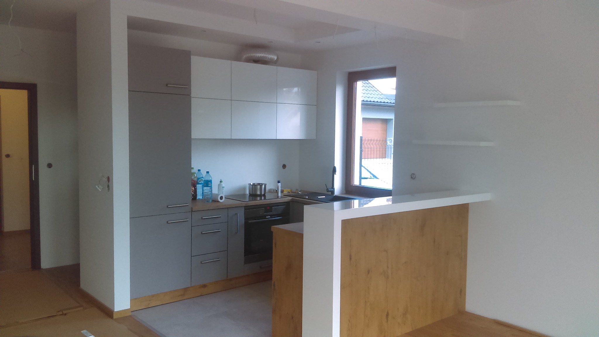 Nowoczesna kuchnia w trakcie montażu: szare dolne szafki z białymi górnymi, drewniany blat-barek z białym wykończeniem, widoczne niedokończone prace wykończeniowe.