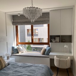 Stolarz Warszawa - Jasna sypialnia z zabudową okienną z siedziskiem, biurkiem i szafkami, okno z drewnianą ramą i szarą roletą rzymską, ozdobny żyrandol.