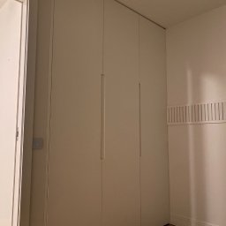 Stolarz Warszawa - Biała, czterodrzwiowa szafa wnękowa z minimalistycznymi uchwytami, zamontowana od podłogi do sufitu, widoczna w rogu pokoju z drewnianą podłogą i grzejnikiem listwowym.