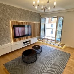 Stolarz Warszawa - Nowoczesny salon z drewnianą ścianą TV, białymi szafkami RTV, dwoma okrągłymi stolikami kawowymi i szarym dywanem w zygzaki, oświetlony designerskim żyrandolem i listwami LED.