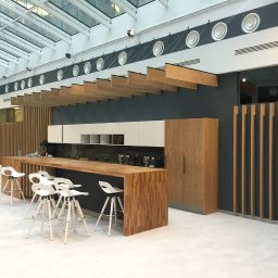 Stolarz Warszawa - Nowoczesna kuchnia z drewnianym blatem i białymi szafkami pod przeszklonym dachem, uzupełniona białymi hokerami i drewnianymi panelami dekoracyjnymi.