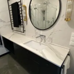 Nowoczesna łazienka z czarną szafką pod umywalką z blatem imitującym marmur, okrągłe lustro z czarną ramą, grzejnik drabinkowy na ręczniki i dekoracyjne złote kinkiety po bokach lustra, marmurowe...