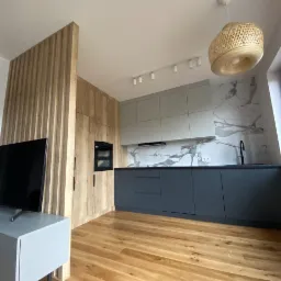 Nowoczesna kuchnia z szarymi szafkami, drewnianymi elementami i parkietem, oświetlona lampą z wikliny i listwą LED, widok na marmurowy backsplash.