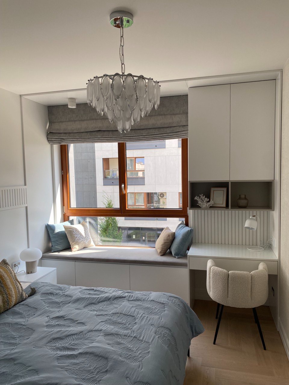 Jasna sypialnia z zabudową okienną z siedziskiem, biurkiem i szafkami, okno z drewnianą ramą i szarą roletą rzymską, ozdobny żyrandol.