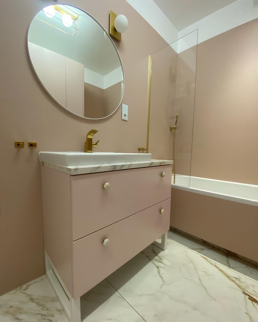 Różowa łazienka z szafką pod umywalkę z marmurowym blatem, złotą armaturą i owalnym lustrem z podświetleniem LED.