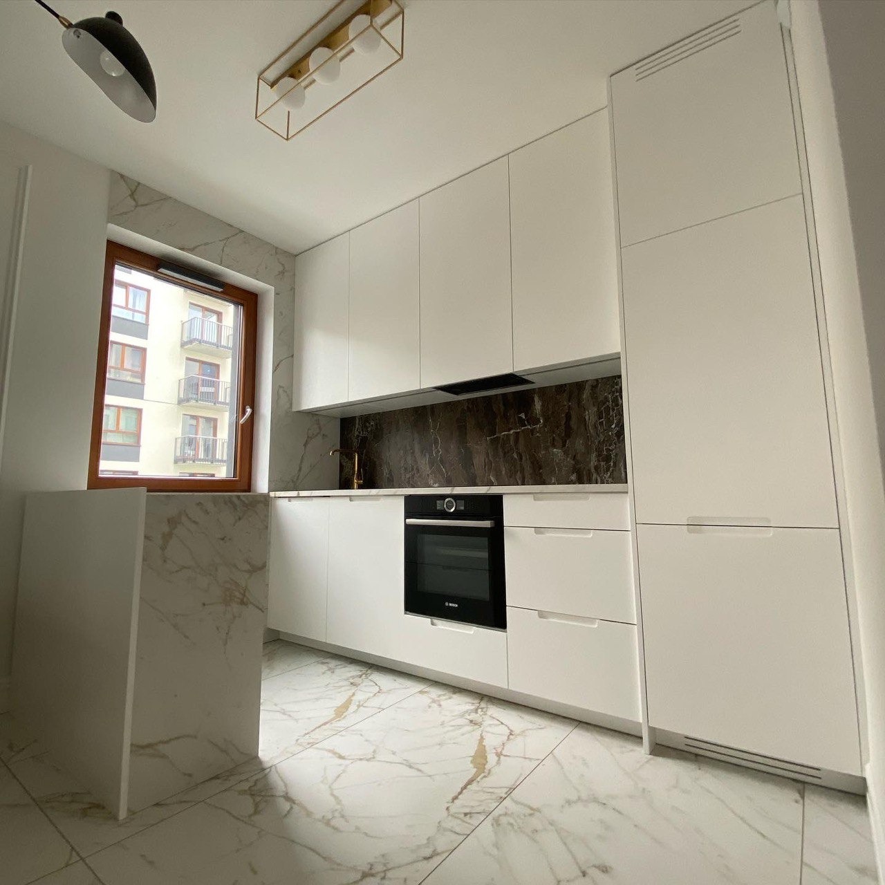 Nowoczesna biała kuchnia na wymiar z marmurowym blatem, brązowym panelem nad blatem, złotą armaturą i lampą sufitową, widok na budynek mieszkalny przez okno.