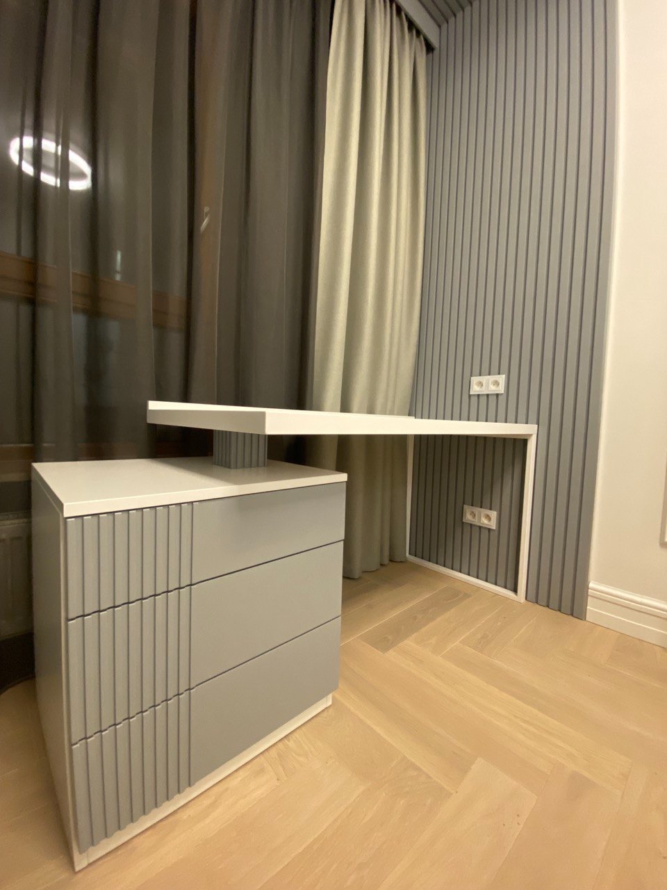 Nowoczesne biurko z szufladami w odcieniach szarości i bieli, z blatem roboczym sięgającym ściany z dekoracyjnymi panelami, na podłodze z parkietu ułożonego w jodełkę.