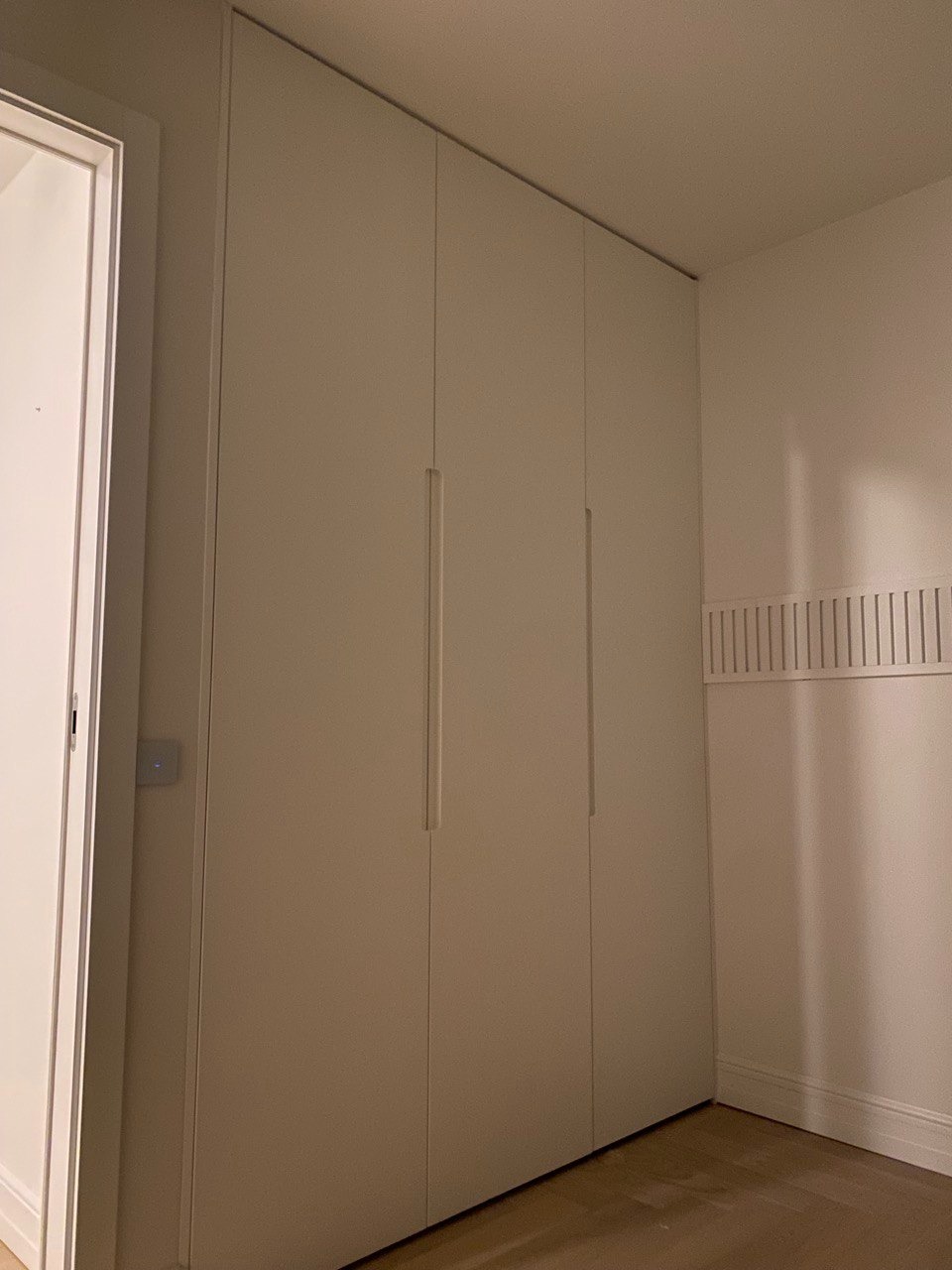 Biała, czterodrzwiowa szafa wnękowa z minimalistycznymi uchwytami, zamontowana od podłogi do sufitu, widoczna w rogu pokoju z drewnianą podłogą i grzejnikiem listwowym.