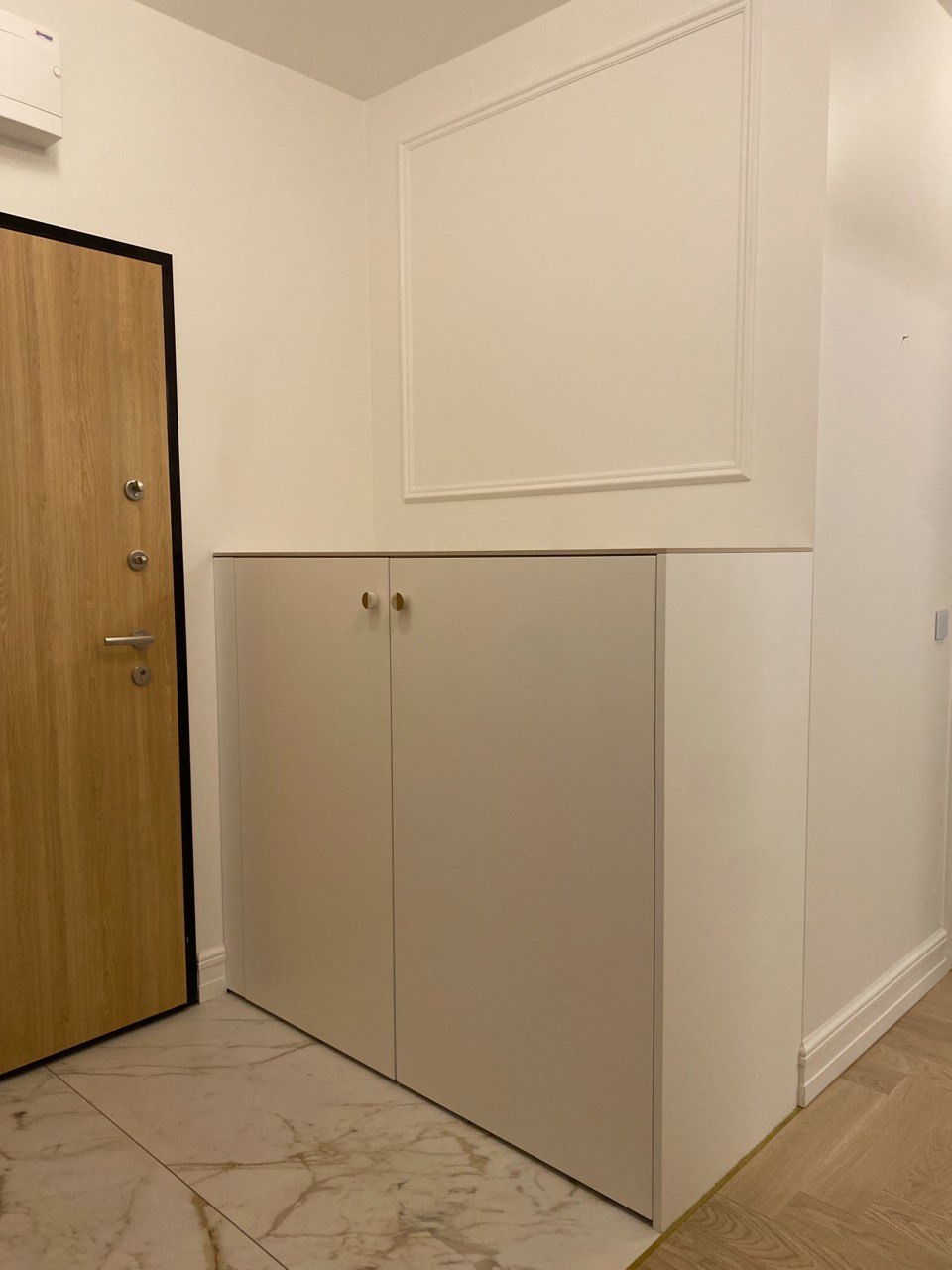 Biała szafka z dwoma drzwiami na złotych uchwytach, stojąca w przedpokoju przy drewnianych drzwiach wejściowych i ścianie z dekoracyjną ramą.