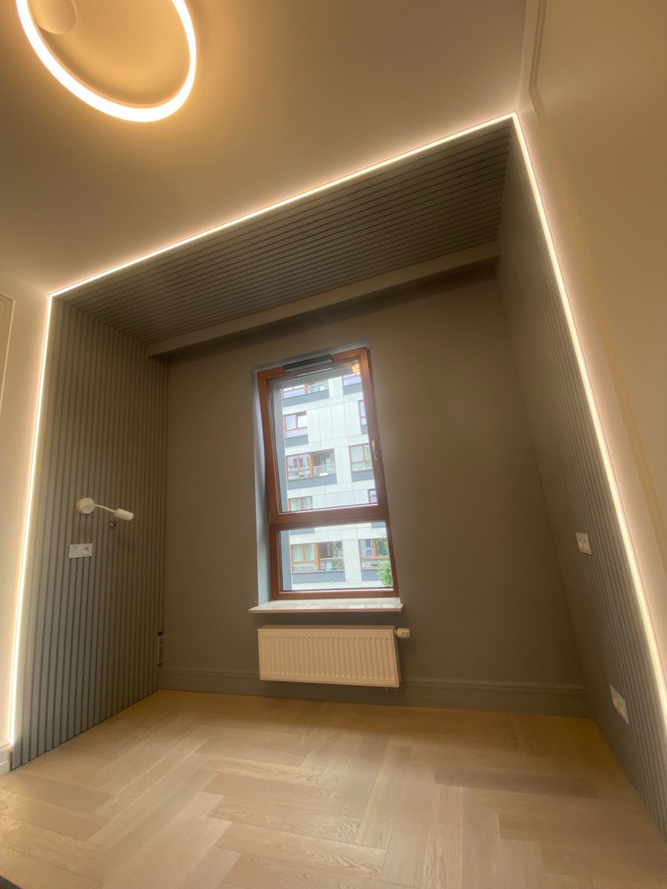 Pusta wnęka z oknem w szarym pokoju, oświetlenie LED wzdłuż krawędzi sufitu i ścian, parkiet w jodełkę na podłodze, grzejnik pod oknem.