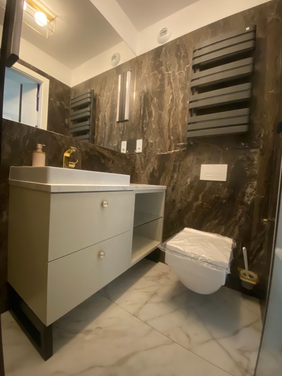 Nowoczesna łazienka z marmurowymi ścianami, białą szafką pod umywalkę z złotymi uchwytami, złotą baterią, grzejnikiem drabinkowym i toaletą zabezpieczoną folią ochronną.
