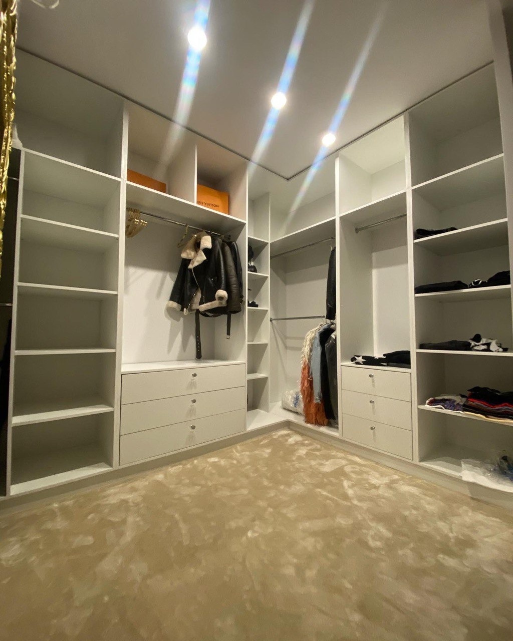 Przestronna garderoba z białymi półkami i szufladami, ubrania wiszące na wieszakach, beżowy dywan na podłodze, oświetlenie punktowe w suficie.