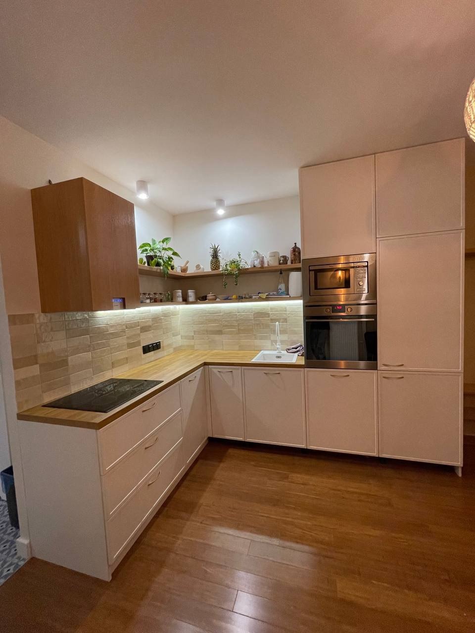 Nowoczesna kuchnia w kształcie litery L z białymi szafkami, drewnianym blatem i płytą indukcyjną, oświetlona ciepłym światłem, z dekoracyjnymi roślinami na otwartej półce.