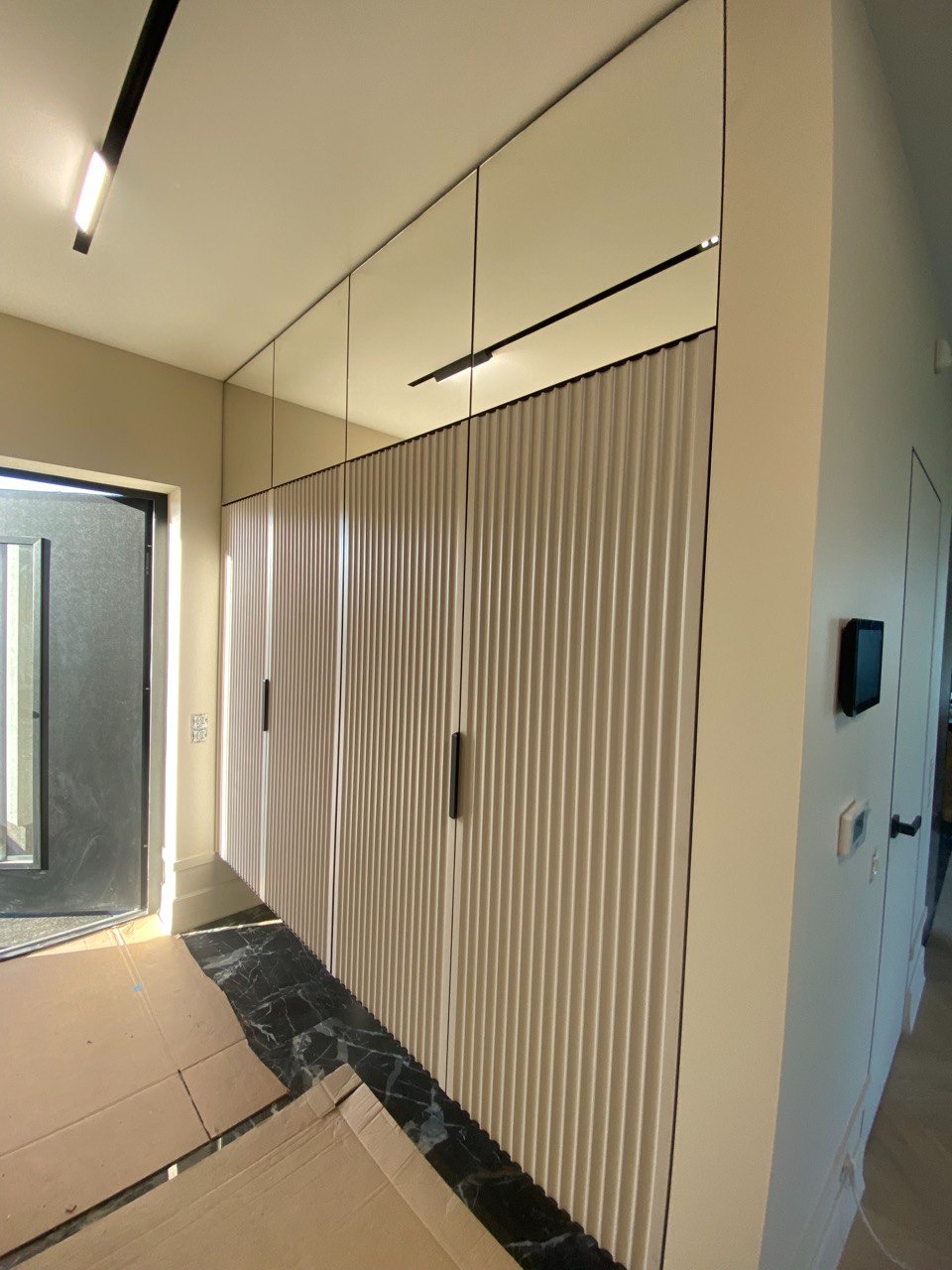 Długa szafa z ryflowanymi drzwiami w przedpokoju, czarne uchwyty i oświetlenie szynowe, marmurowa podłoga, zabezpieczona kartonem.