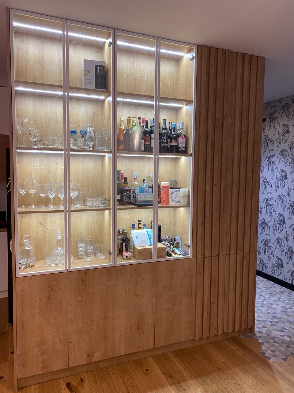Drewniana zabudowa wnęki z przeszklonymi szafkami z oświetleniem LED, wypełniona szkłem i butelkami, wykonana na wymiar w Lomiankach, z dekoracyjnym panelem ściennym.