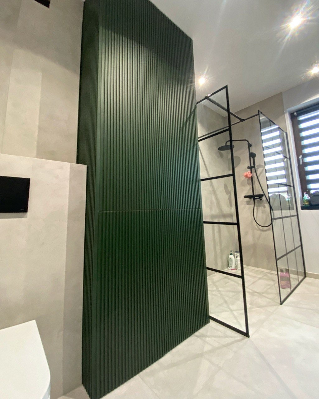 Nowoczesna łazienka z kabiną prysznicową typu walk-in, oddzielona zielonym, pionowo żłobionym panelem dekoracyjnym, widoczne białe sanitariaty i okno z żaluzjami.