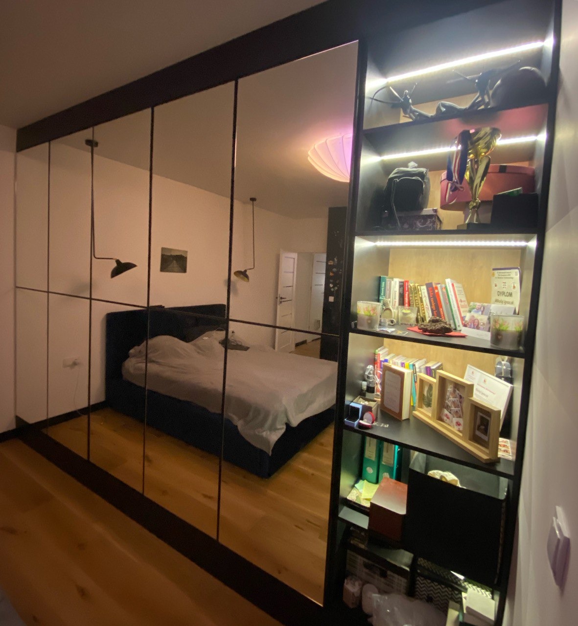 Duża szafa z lustrzanymi drzwiami przesuwnymi, odzwierciedlająca sypialnię z łóżkiem i lampami wiszącymi, z boku regał z podświetlanymi półkami, na których znajdują się książki, dyplomy i dekoracje.