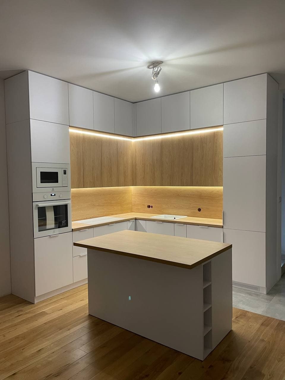 Nowoczesna kuchnia z białymi szafkami i drewnianymi frontami podświetlonymi LED, wyspa kuchenna z jasnym blatem i otwartymi półkami, parkiet na podłodze.