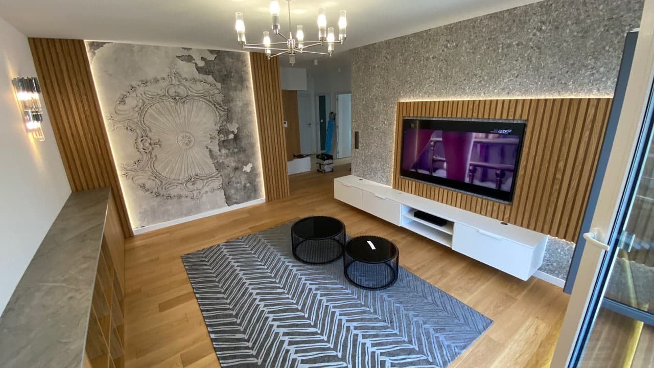 Nowoczesny salon z drewnianymi panelami ściennymi, szarą tapetą z ornamentem, niską białą szafką RTV oraz dwoma okrągłymi stolikami kawowymi na wzorzystym dywanie.