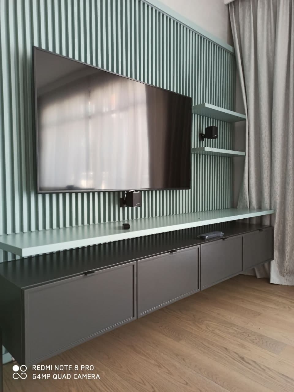 Nowoczesna szafka RTV w kolorze grafitowym z minimalistycznymi uchwytami, zawieszona pod telewizorem na tle ściany z pionowymi panelami w odcieniach zieleni i szarości, uzupełniona półkami i zasłoną.