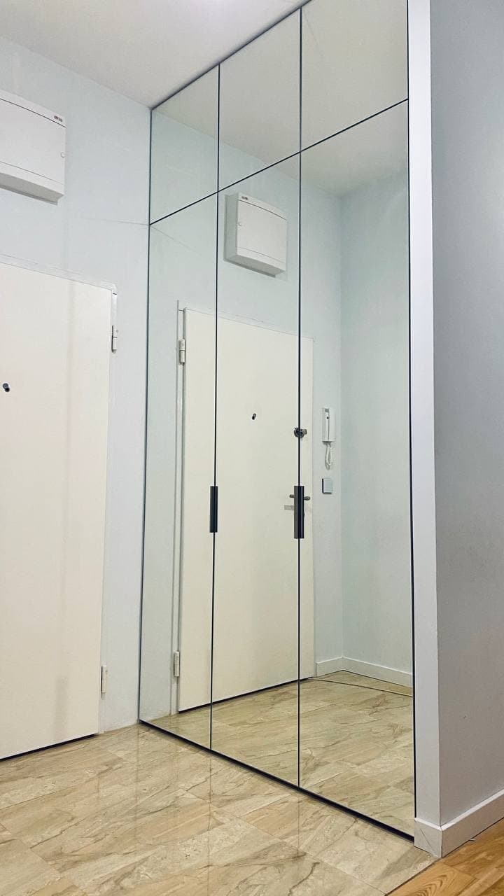 Wysoka szafa z lustrzanymi drzwiami przesuwnymi, z czarnymi, pionowymi listwami dzielącymi, odbijająca podłogę z płytek imitujących marmur i białe drzwi wejściowe.