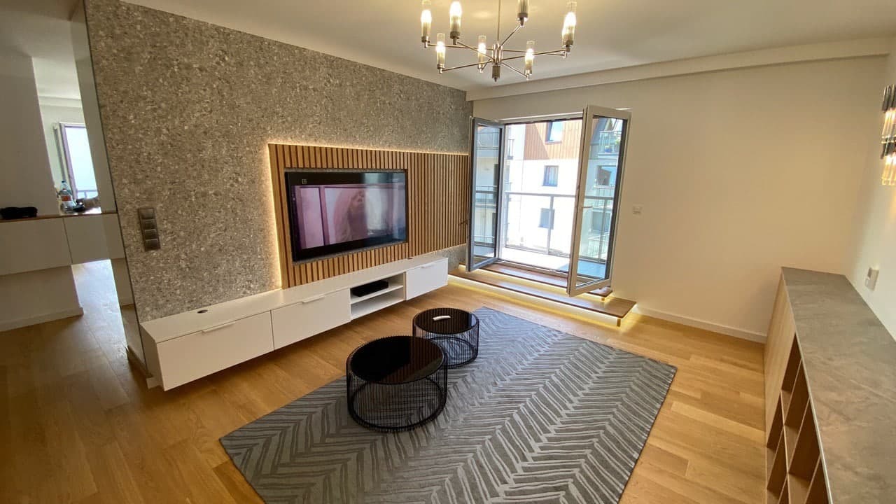 Nowoczesny salon z drewnianą ścianą TV, białymi szafkami RTV, dwoma okrągłymi stolikami kawowymi i szarym dywanem w zygzaki, oświetlony designerskim żyrandolem i listwami LED.