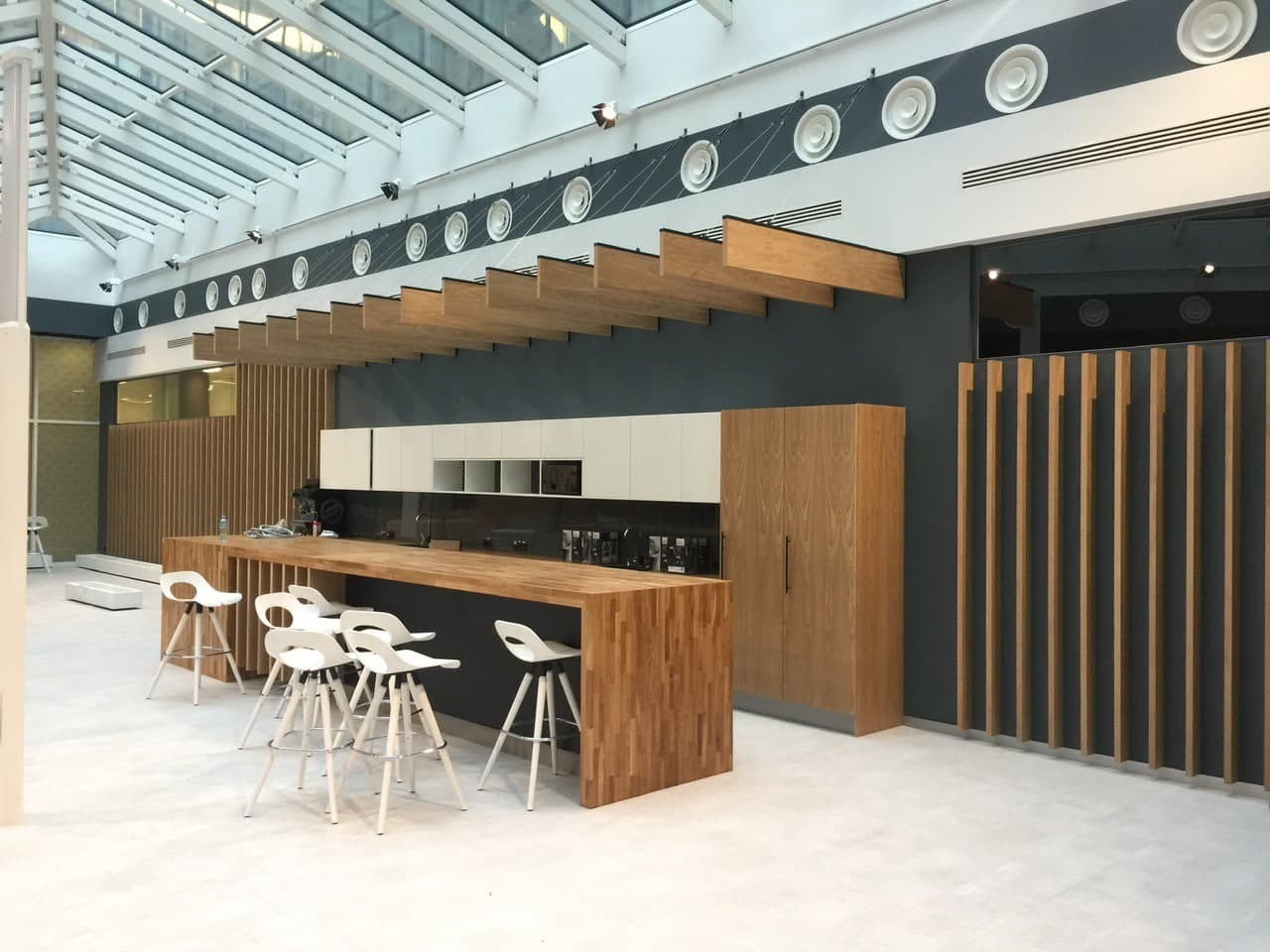 Nowoczesna kuchnia z drewnianym blatem i białymi szafkami pod przeszklonym dachem, uzupełniona białymi hokerami i drewnianymi panelami dekoracyjnymi.