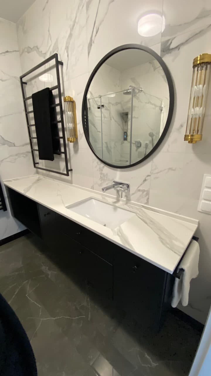 Nowoczesna łazienka z czarną szafką pod umywalką z blatem imitującym marmur, okrągłe lustro z czarną ramą, grzejnik drabinkowy na ręczniki i dekoracyjne złote kinkiety po bokach lustra, marmurowe...