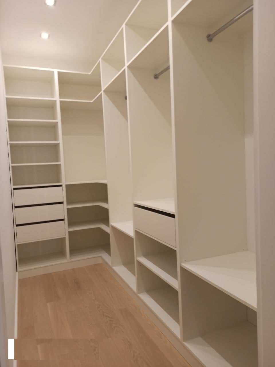 Wnętrze białej, przestronnej garderoby wnękowej z półkami, szufladami z czarnymi uchwytami i drążkami na ubrania, na jasnej drewnianej podłodze.