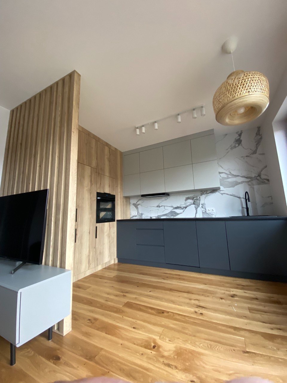 Nowoczesna kuchnia z szarymi szafkami, drewnianymi elementami i parkietem, oświetlona lampą z wikliny i listwą LED, widok na marmurowy backsplash.