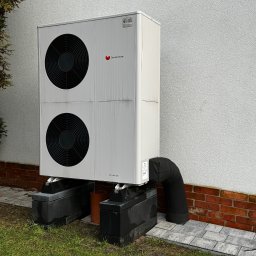 Instalsan Sp. z o.o. - Biała pompa ciepła Saunier Duval GeniaAir Split, zamontowana na betonowych blokach przy ścianie budynku z ceglaną podmurówką i trawnikiem.