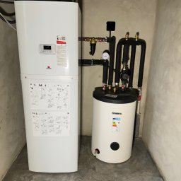 Instalsan Sp. z o.o. - Biała pompa ciepła z zasobnikiem Kospel SVK-100 w kotłowni. Widoczne rury, manometr i instrukcje montażu na obudowie. Szare ściany i podłoga.