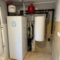 Instalsan Sp. z o.o. - Zmontowana instalacja z pompą ciepła LG Therma V, zbiornikiem Galmet i rurami w piwnicy. Widoczne zawory, manometry i elementy mocujące. Beżowe płytki na ścianach i podłodze.