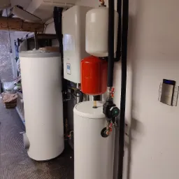 Kompletna instalacja grzewcza z białym zbiornikiem wody, czerwonym naczyniem przeponowym, czarnymi rurami, pompą obiegową i panelem sterowania na ścianie.