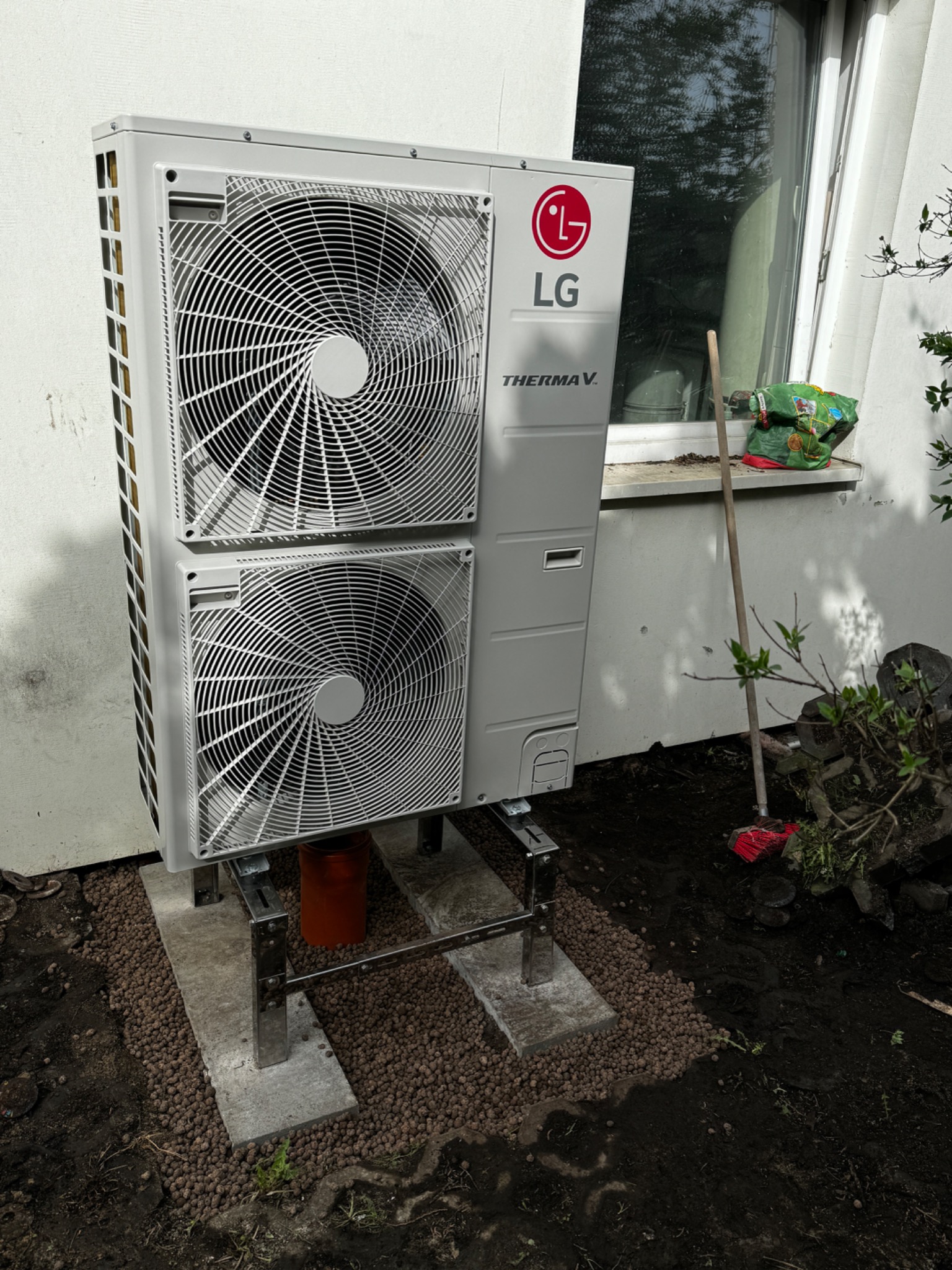 Zewnętrzna jednostka pompy ciepła LG Therma V, zamontowana na betonowych podstawach z podsypką z keramzytu, przy białej ścianie budynku z oknem.