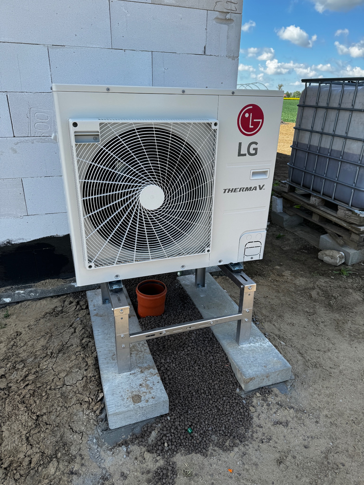 Biała pompa ciepła LG Therma V, zamontowana na betonowej podstawie z metalową ramą, na tle ściany z bloczków i zbiornika na wodę. Widoczne pole w tle.