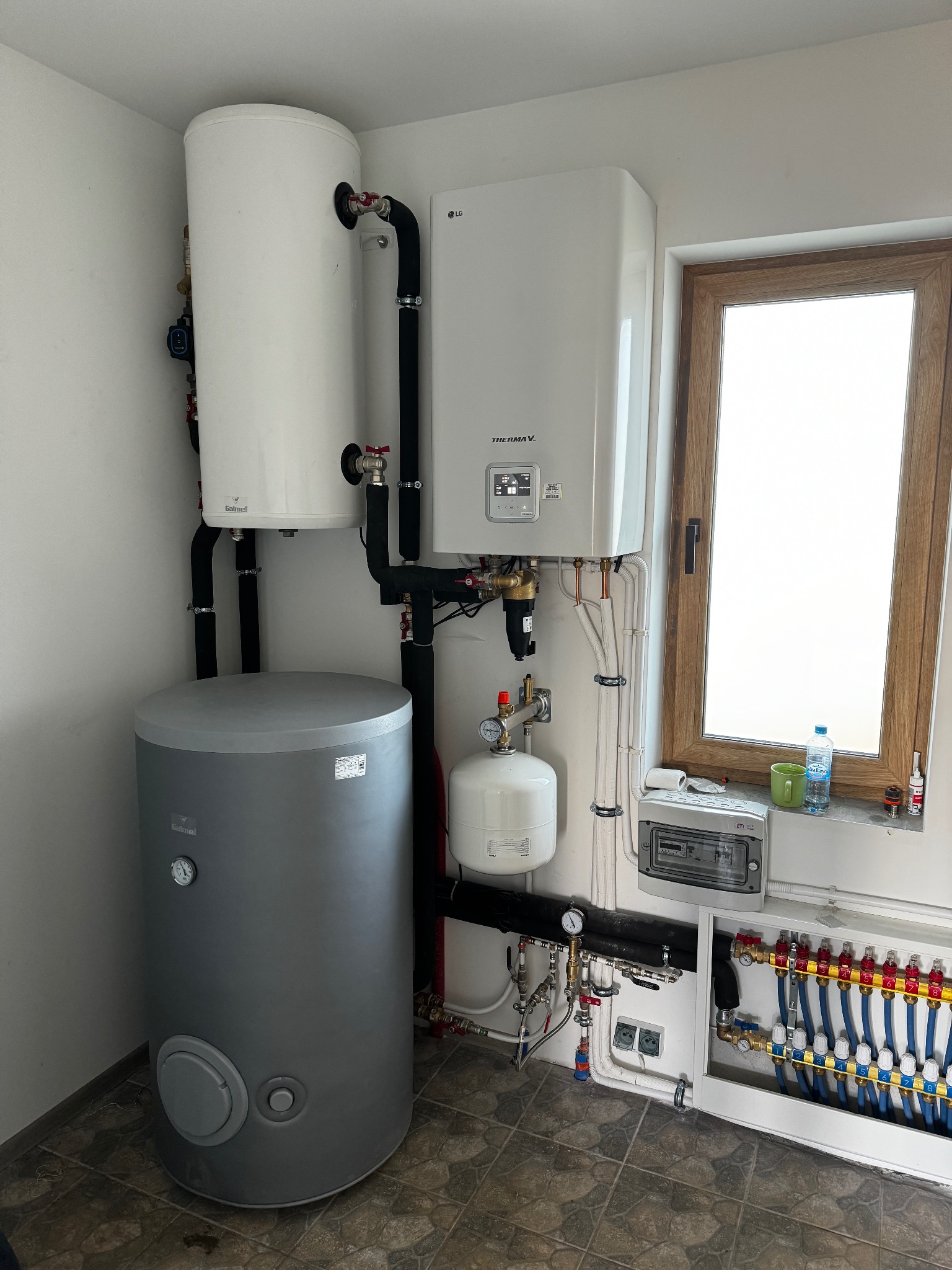 Kompletna instalacja grzewcza z piecem gazowym LG Therma V, zbiornikami Galmet i rozdzielaczem ogrzewania podłogowego, zamontowana w pomieszczeniu z oknem.
