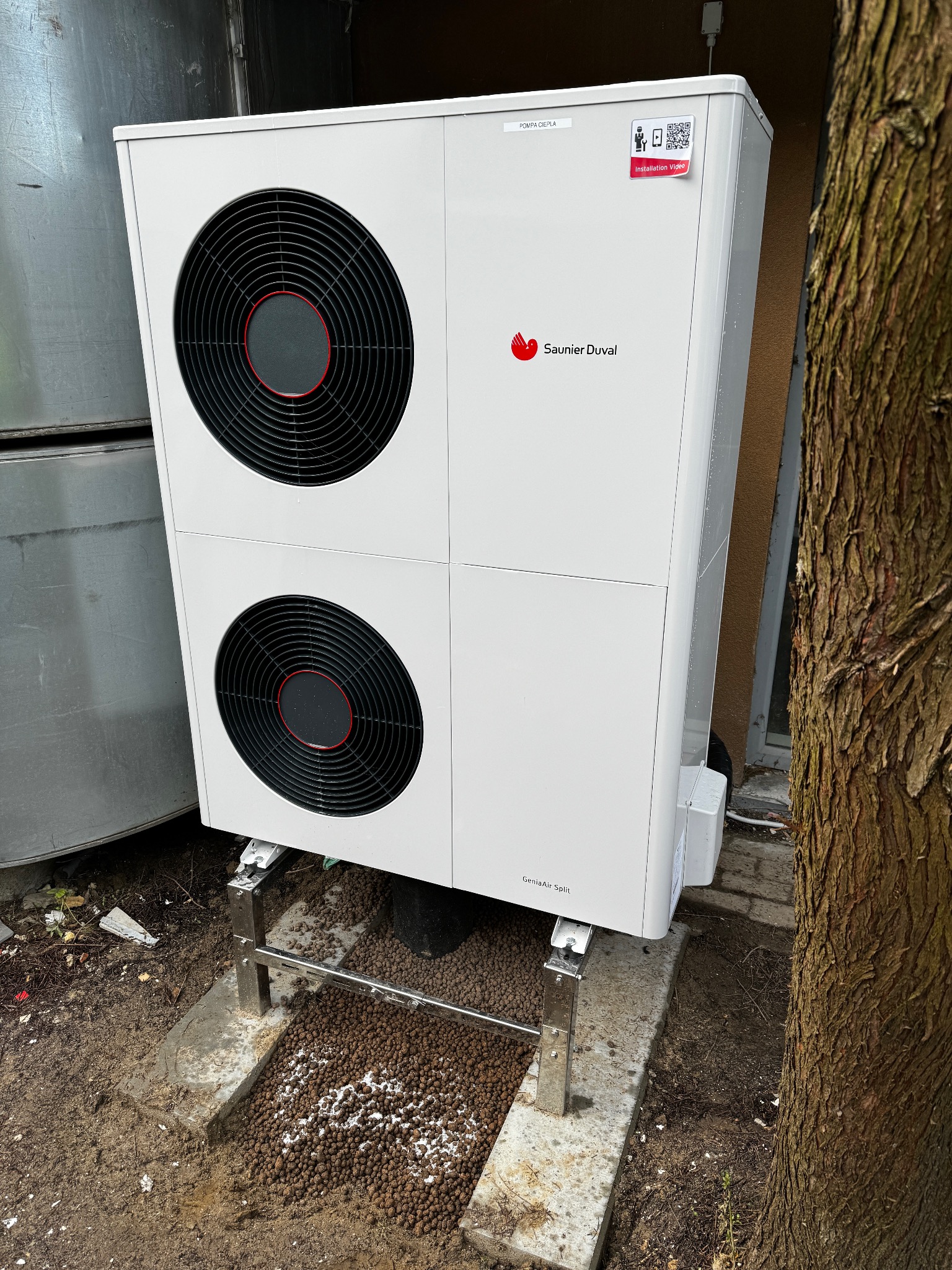 Biała pompa ciepła Saunier Duval GeniaAir Split, zamontowana na metalowej konstrukcji, z widocznymi dwoma wentylatorami i naklejką z kodem QR, obok drzewa.